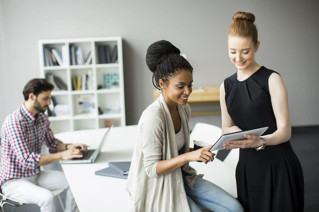 Three colleagues in a modern office: two women discuss a tablet while a man works on a laptop in the background.