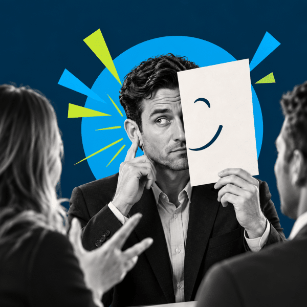 Man in a suit holds a paper with a smiley mask over half of his face during a meeting with colleagues.